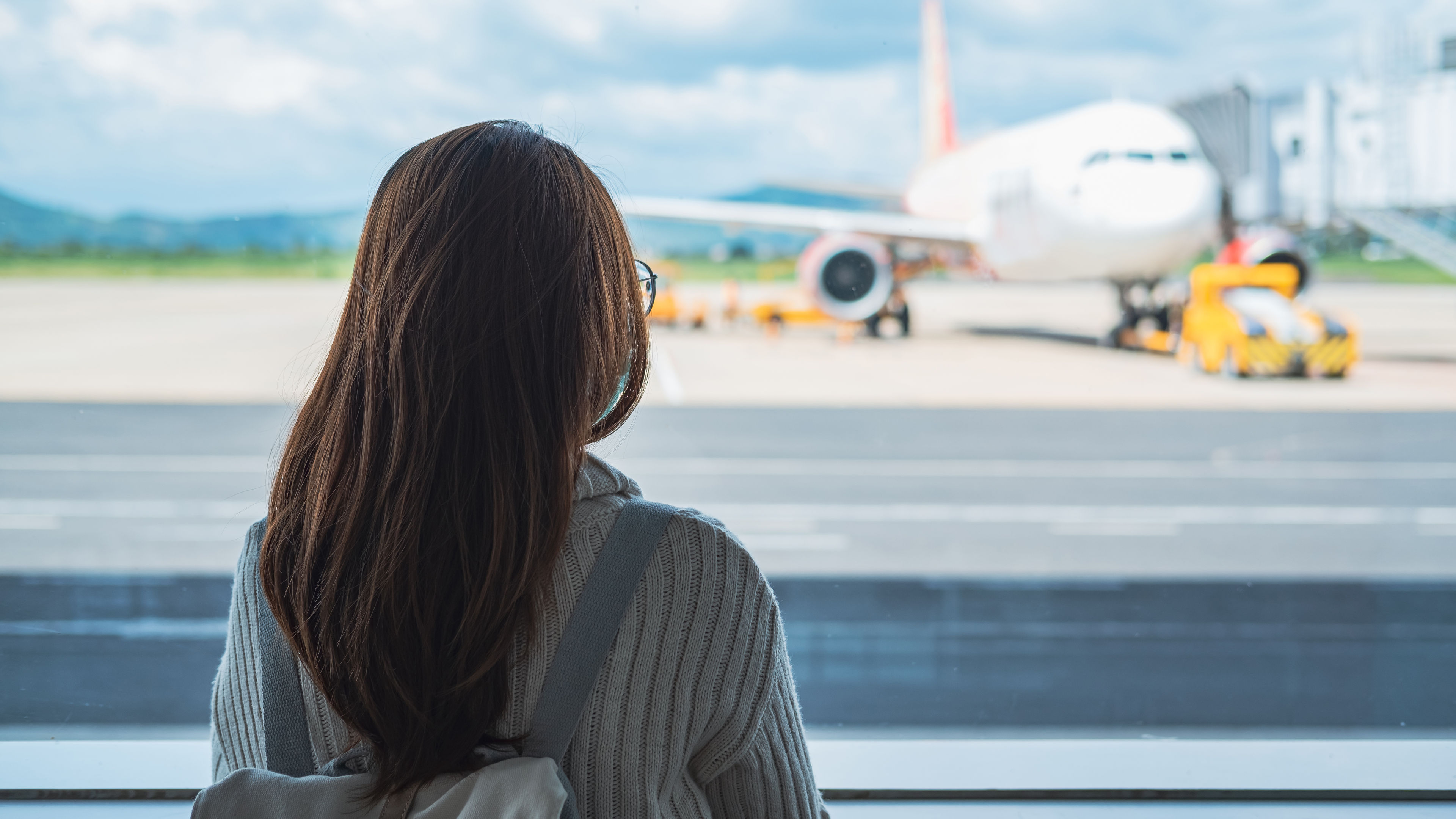 A backpacked traveler leans against an airport window, observing planes on the runway as they prepare for departure.