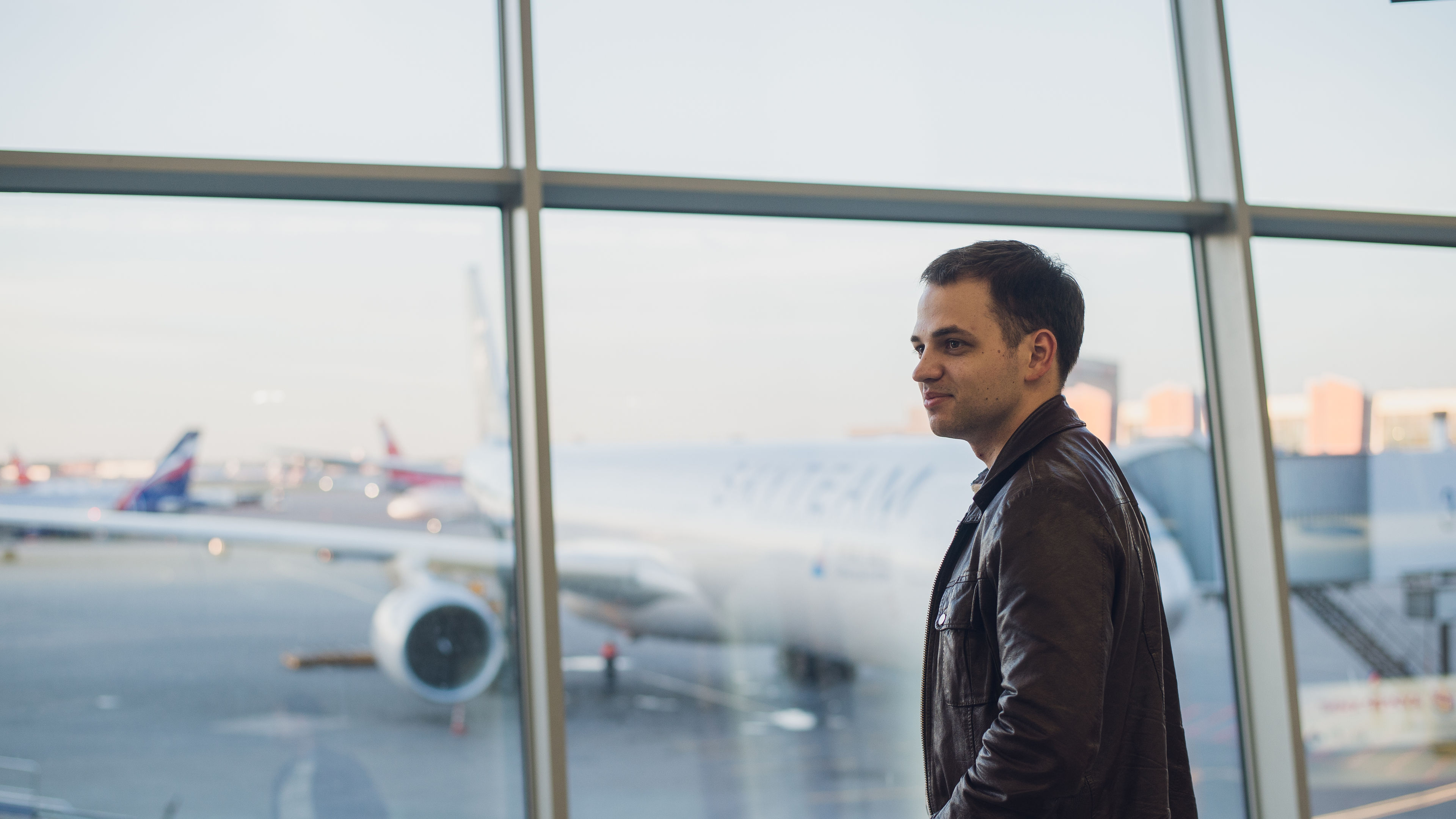 A young man posing near an airport window with planes in the background.