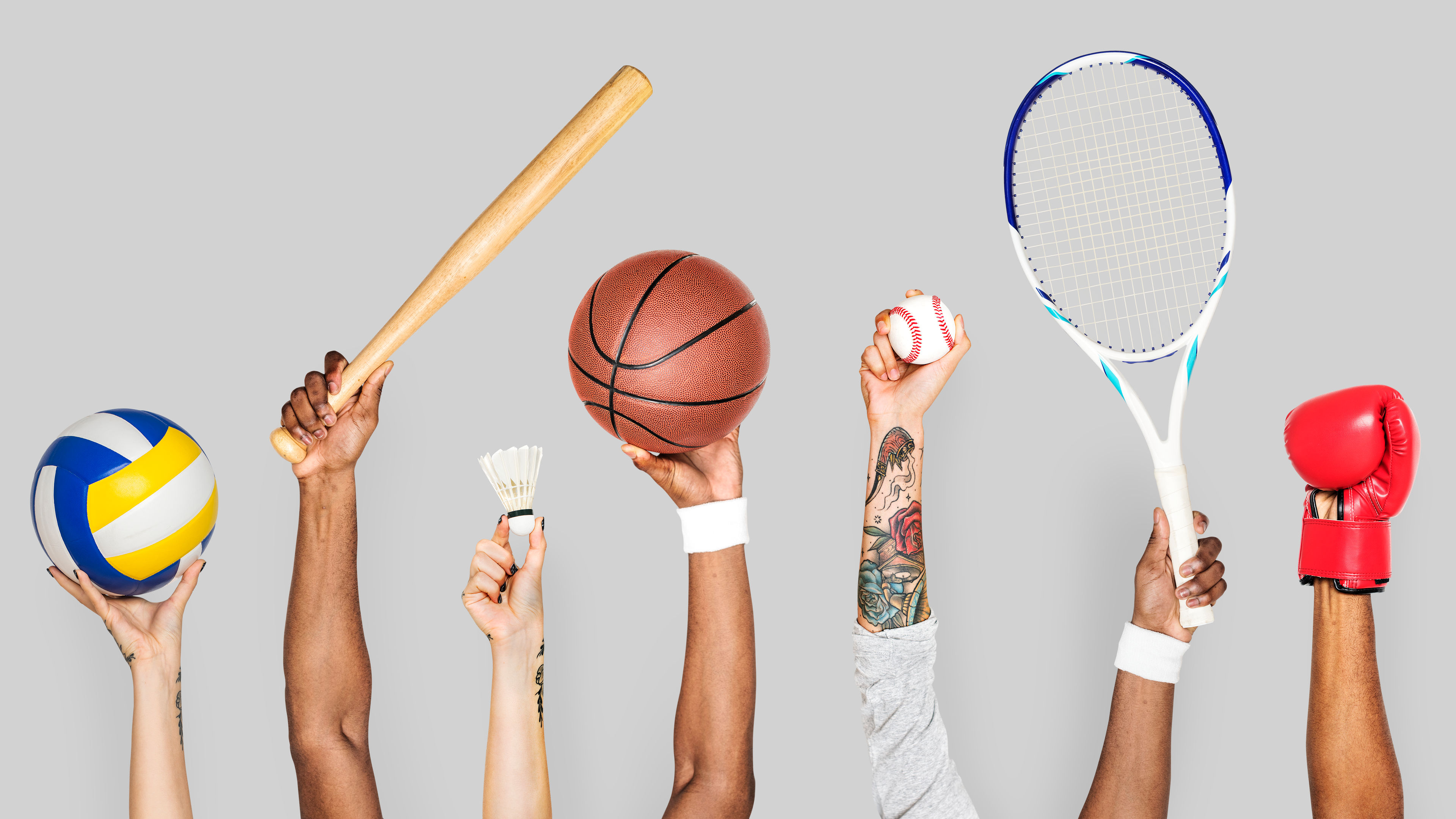 Hands holding a volleyball, bat, shuttlecock, basketball, baseball, racket and boxing glove in grey background.
